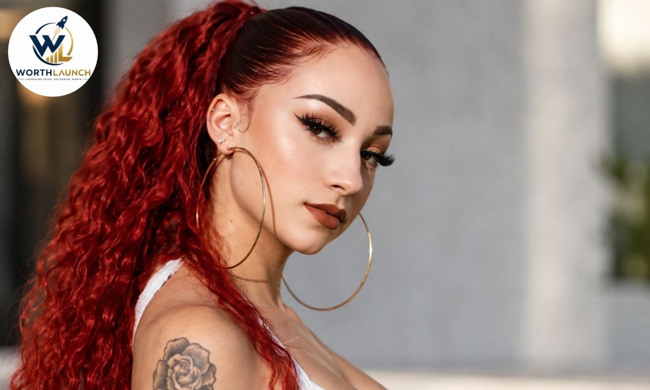 Young woman with long red curly hair, bold makeup, hoop earrings, and a rose shoulder tattoo posing in a professional portrait.