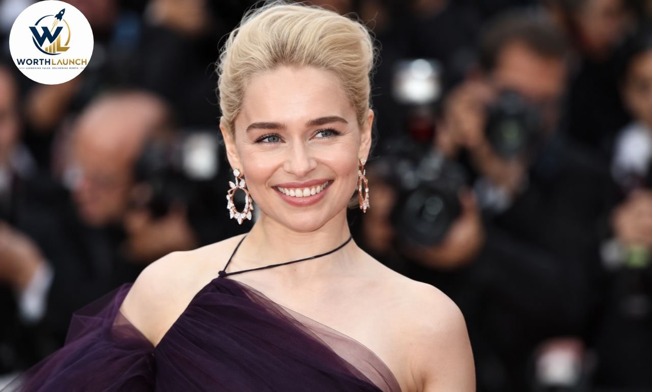 Emilia Clarke smiling on the red carpet in a deep purple one-shoulder gown with elegant earrings.
