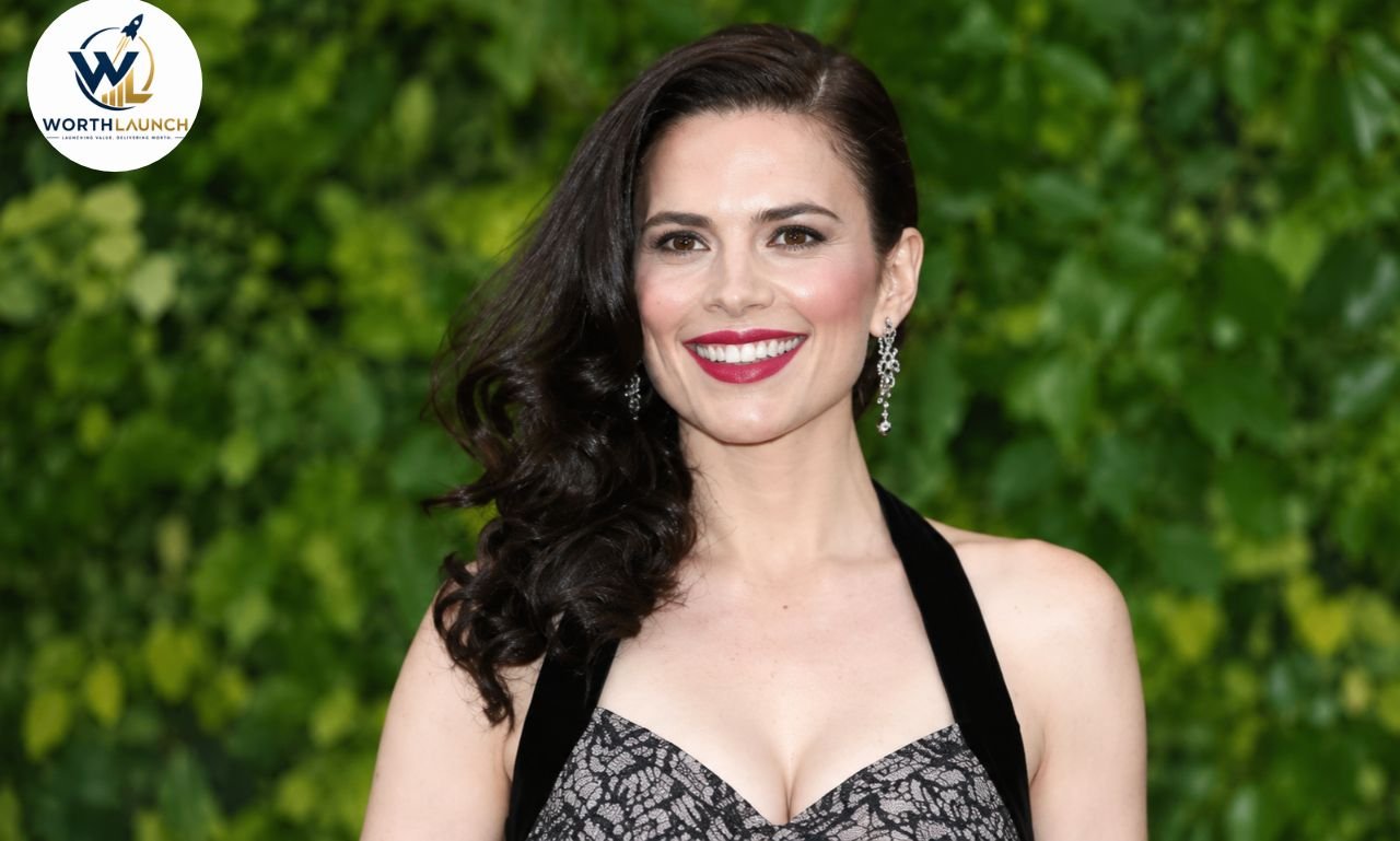Elegant portrait Hayley Atwell in a black lace dress with red lipstick and sparkling earrings against a lush green background.