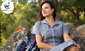 Kathryn Burrhus sitting outdoors on rocks in a natural setting, wearing a gray button-up shirt and looking thoughtfully into the distance.