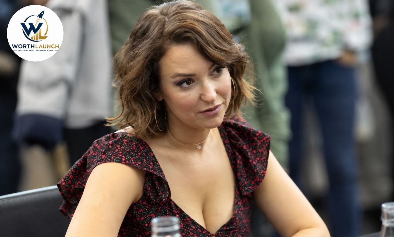 Milana Vayntrub in a black and red floral dress seated at an event, looking attentively to the side.