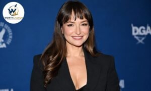 Milana Vayntrub in a black blazer posing at a red-carpet event against a blue backdrop.