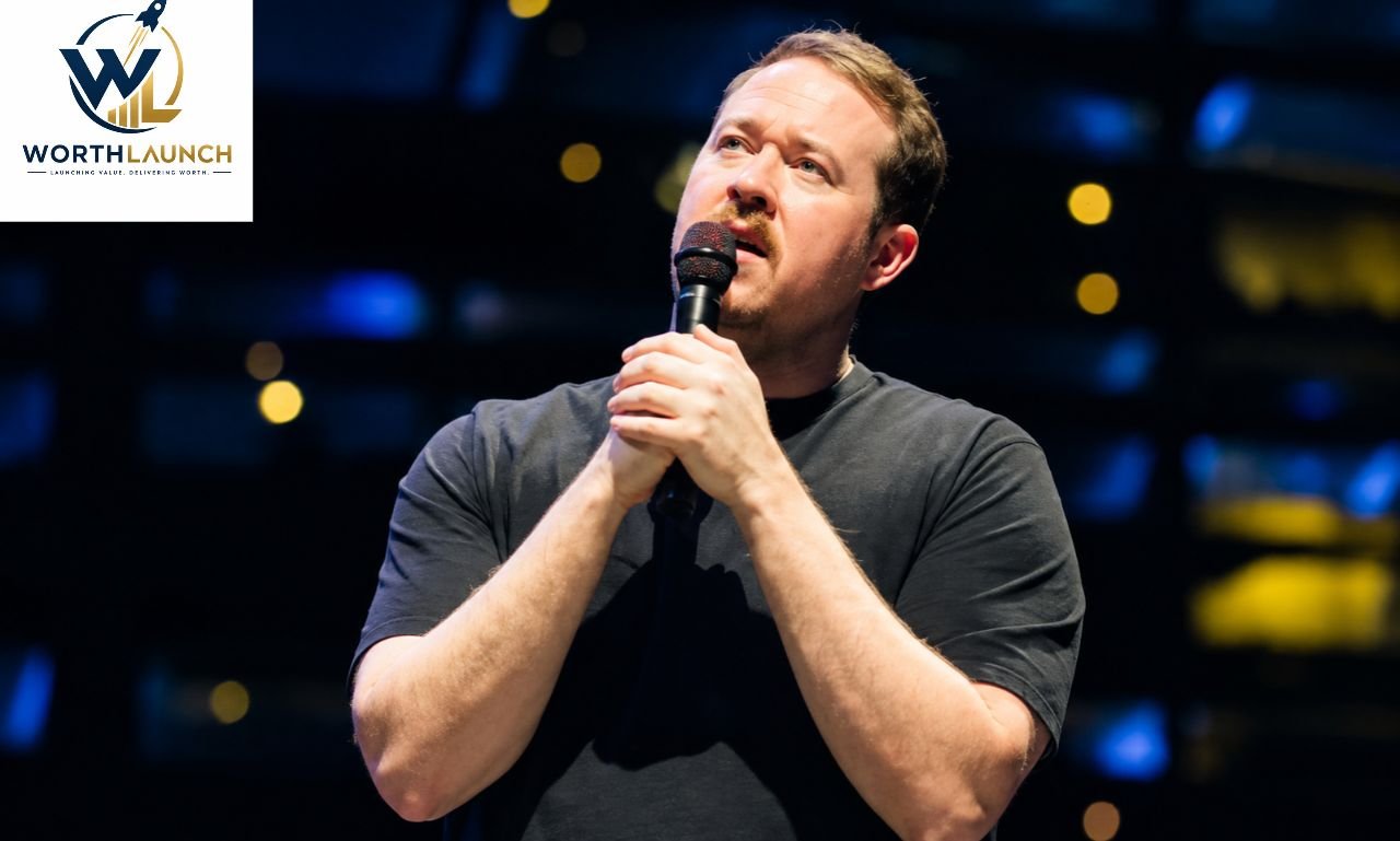 Shane Gillis performing stand-up comedy on stage while holding a microphone under dramatic lighting.
