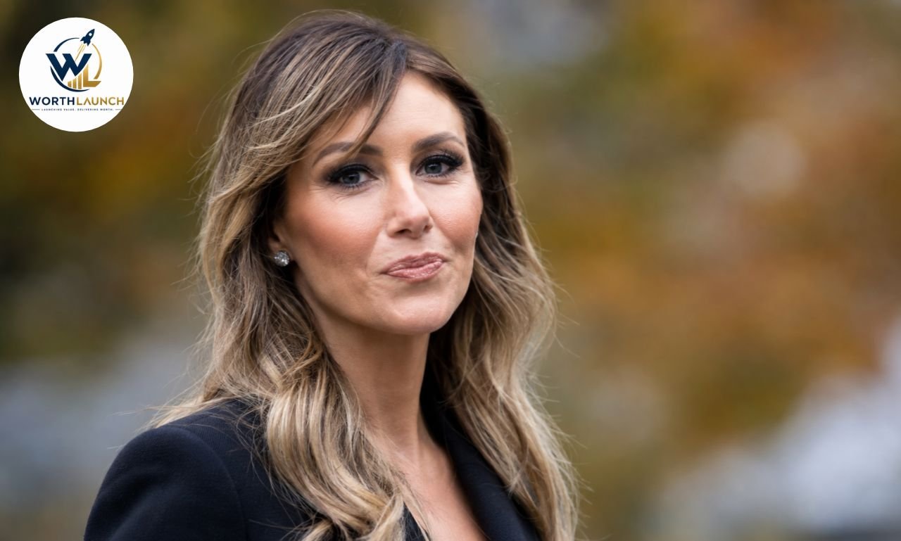 Alina Habba in a black blazer with long wavy hair posing outdoors against a blurred autumn background.