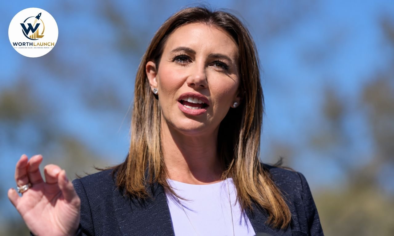 Alina Habba  in a dark blazer speaking outdoors with one hand raised against a blue sky.