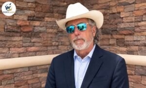 Arch Aplin III wearing a white cowboy hat, blue mirrored sunglasses, and a navy blazer against a stone wall.