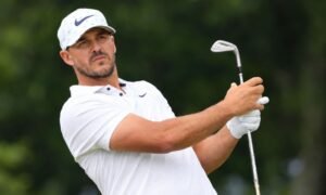 Brooks Koepka playing golf during a swing while wearing a white Nike cap and polo on a golf course.