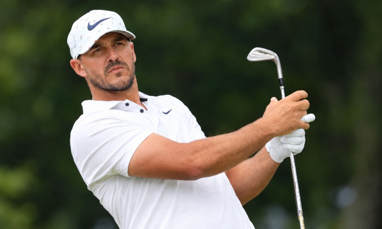 Brooks Koepka playing golf during a swing while wearing a white Nike cap and polo on a golf course.