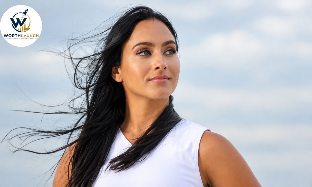 Gabi Butler with long black hair blowing in the wind, wearing a white sleeveless top, gazing calmly to the side against a soft blue-gray sky.