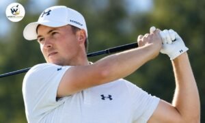 Jordan Spieth playing golf in a white Under Armour shirt and cap during a follow-through shot on a sunny course.