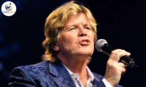 Peter Noone singing into a microphone during a live concert performance.