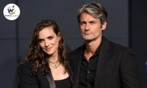 Scott Mackinlay Hahn posing with a woman at a red-carpet event, both dressed in elegant black outfits against a dark background.